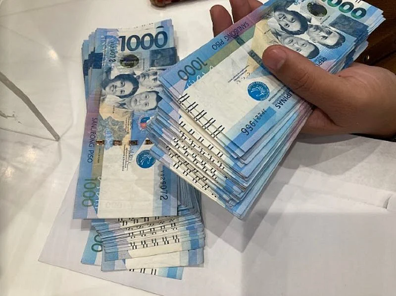 A person holds a Philippine peso banknote against a blurred cityscape background of modern skyscrapers.