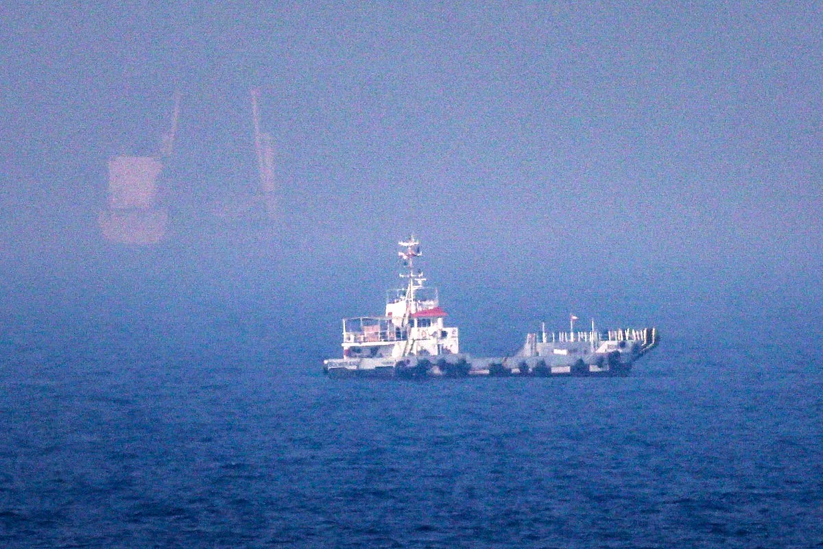 A large container ship navigates deep blue waters near a rocky shoreline under a hazy, bright sky.