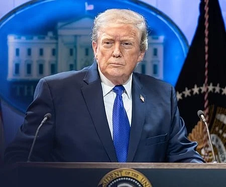 Donald Trump gestures while speaking at a podium against a backdrop of several American flags.