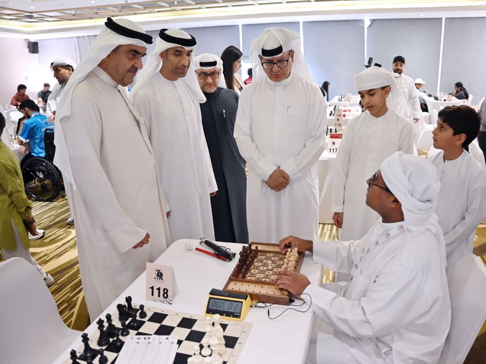 Rows of focused competitors sit at long tables with chessboards during a large-scale tournament in a bright arena.