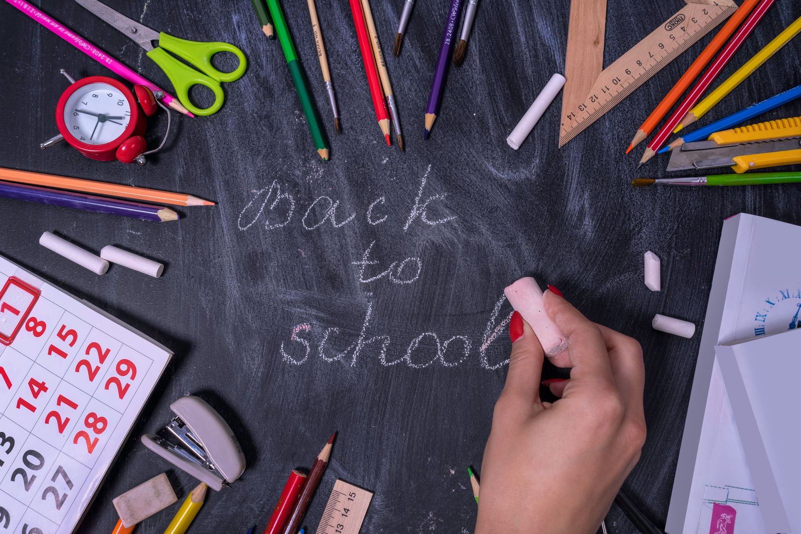 A student in a school uniform sits at a desk with a colorful backpack, notebooks, and pencils in a bright room.