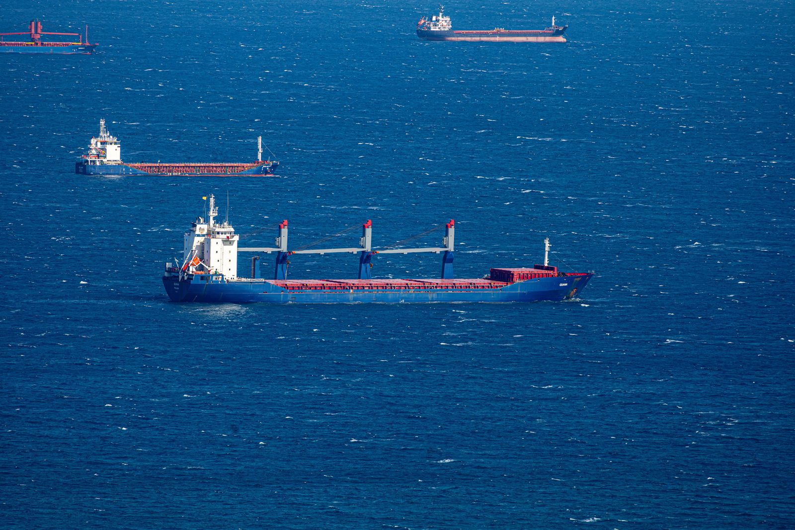 Cargo ships navigate through the narrow Strait of Hormuz between desert coastlines under a clear blue sky.