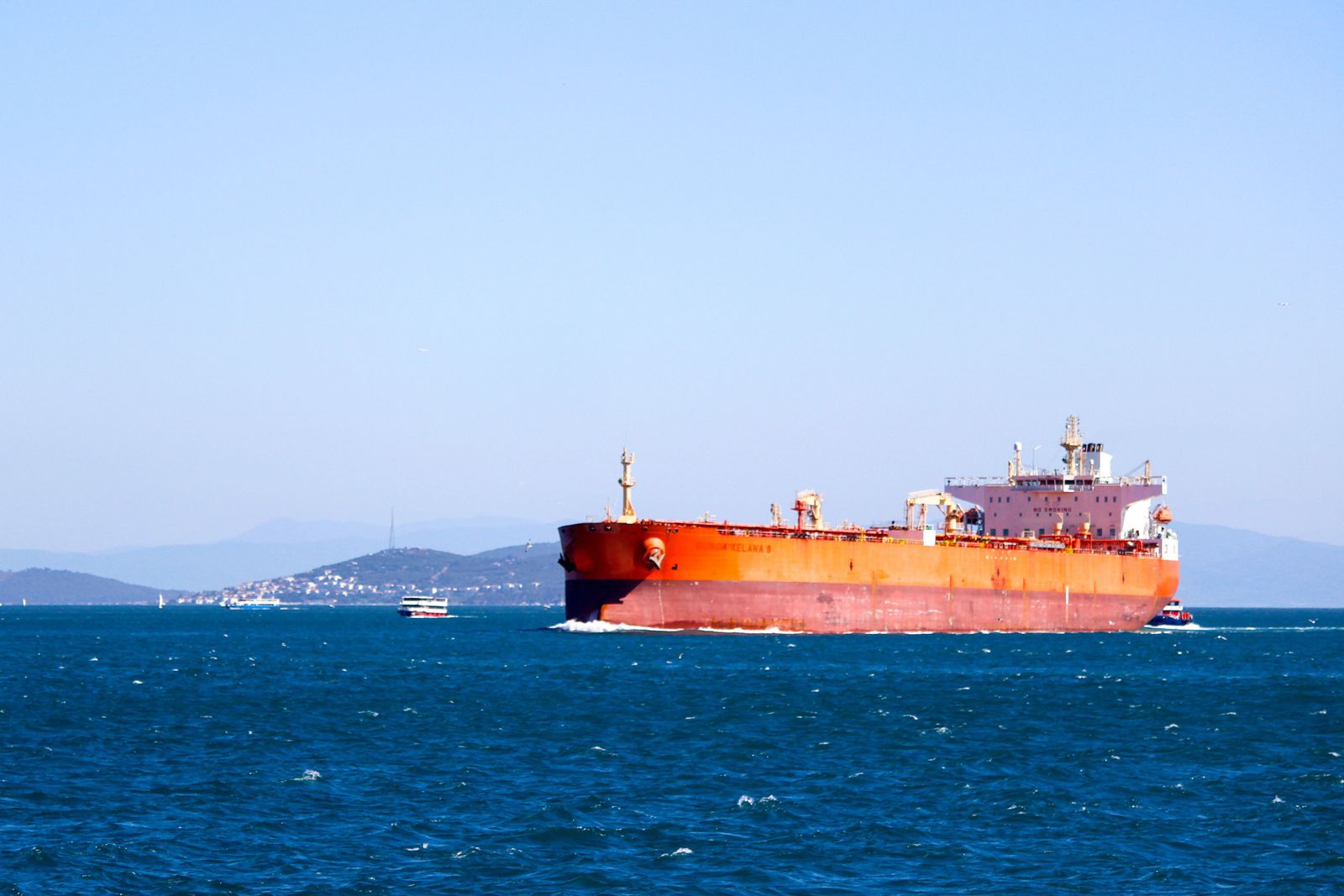 A large oil tanker sails through the narrow blue waters of the Strait of Hormuz framed by arid mountains.