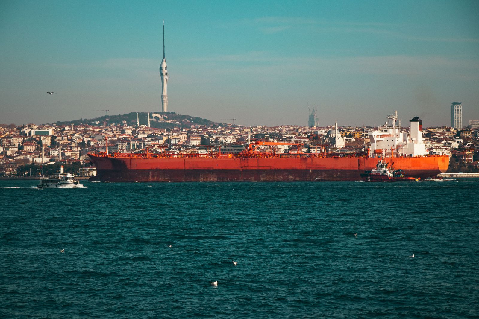 Thick black smoke billows from the deck of a large red and gray tanker ship adrift in blue ocean waters.