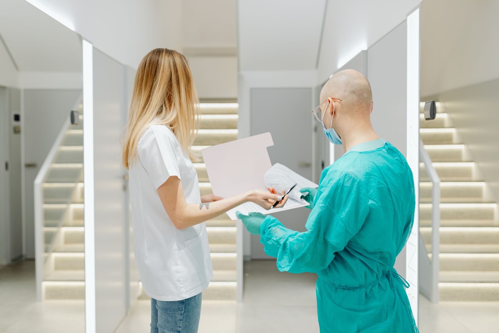 A female doctor wearing a white lab coat and stethoscope smiles while standing in a modern hospital hallway.