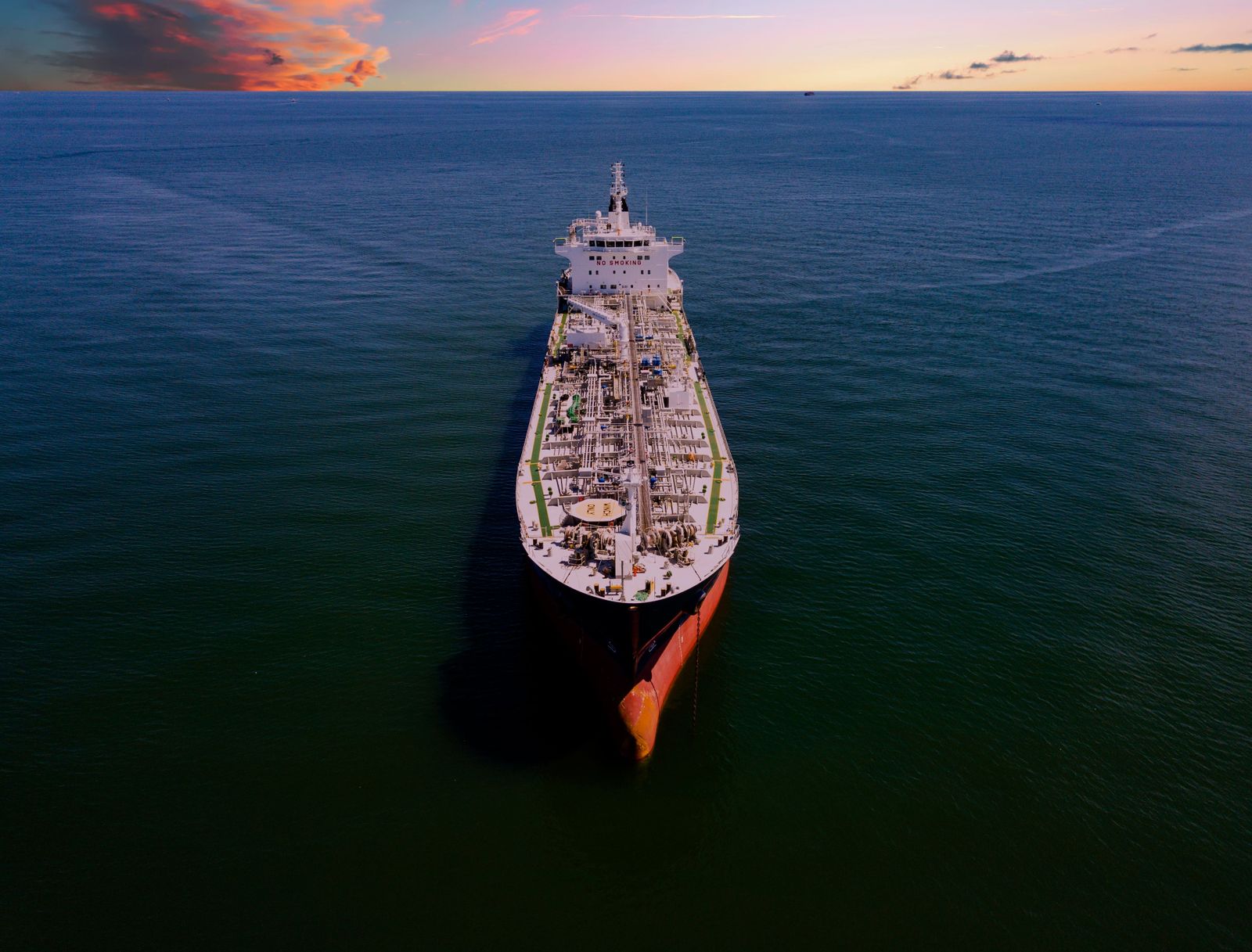 A container ship navigates deep blue waters near a mountainous coastline under a hazy, industrial sky.