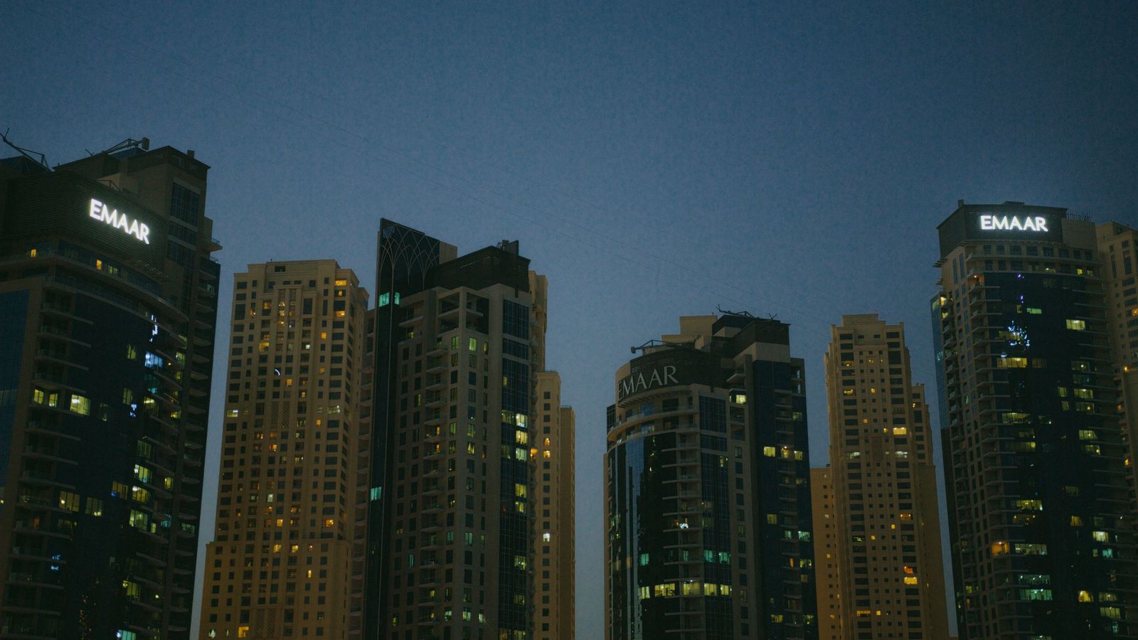 The flag of the United Arab Emirates flutters in front of a modern glass skyscraper in a bright financial district.