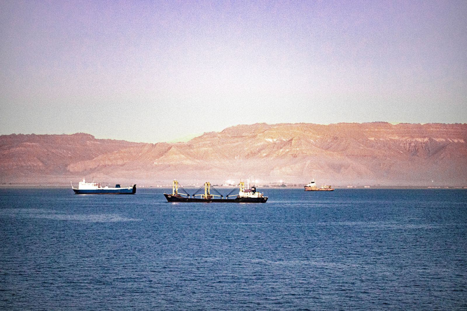 Large oil tankers navigate the blue waters of the Strait of Hormuz near rugged, mountainous desert coastlines.