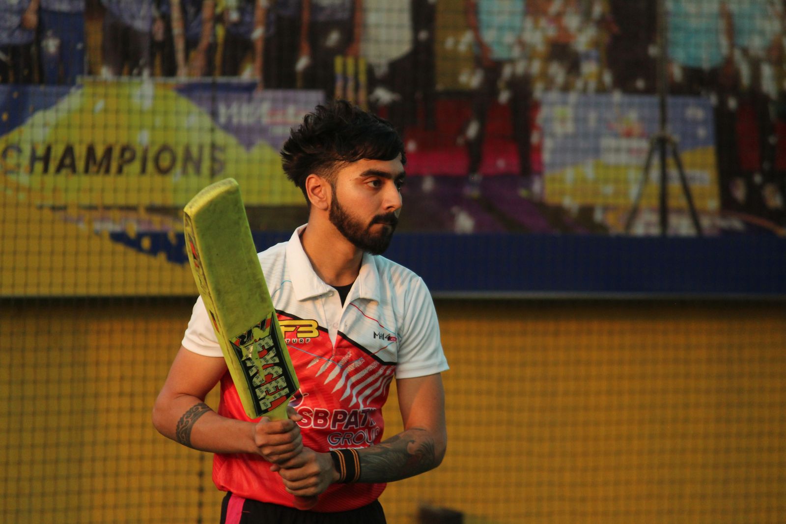 Babar Azam holds a cricket bat while wearing his green Pakistan jersey against a blurred stadium backdrop.