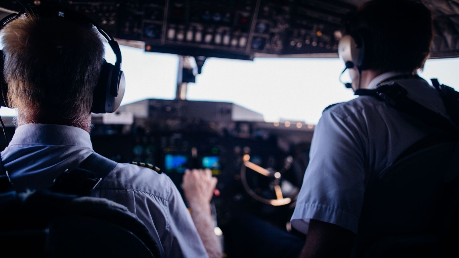 A pilot in a white uniform sits in a flight cockpit surrounded by glowing instrument panels and navigation controls.