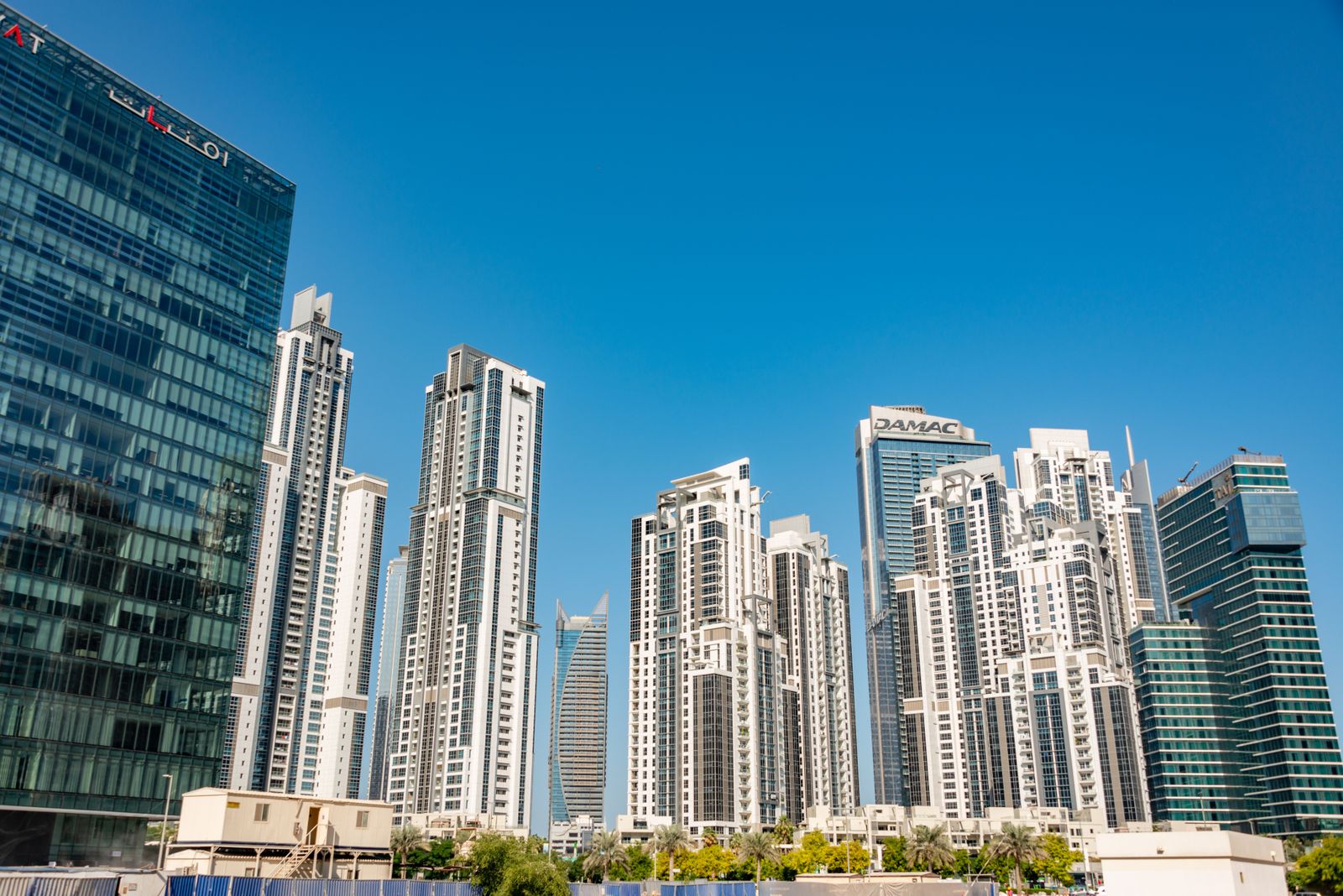 Modern residential skyscrapers tower over a palm-lined street in Dubai under a clear blue sky.
