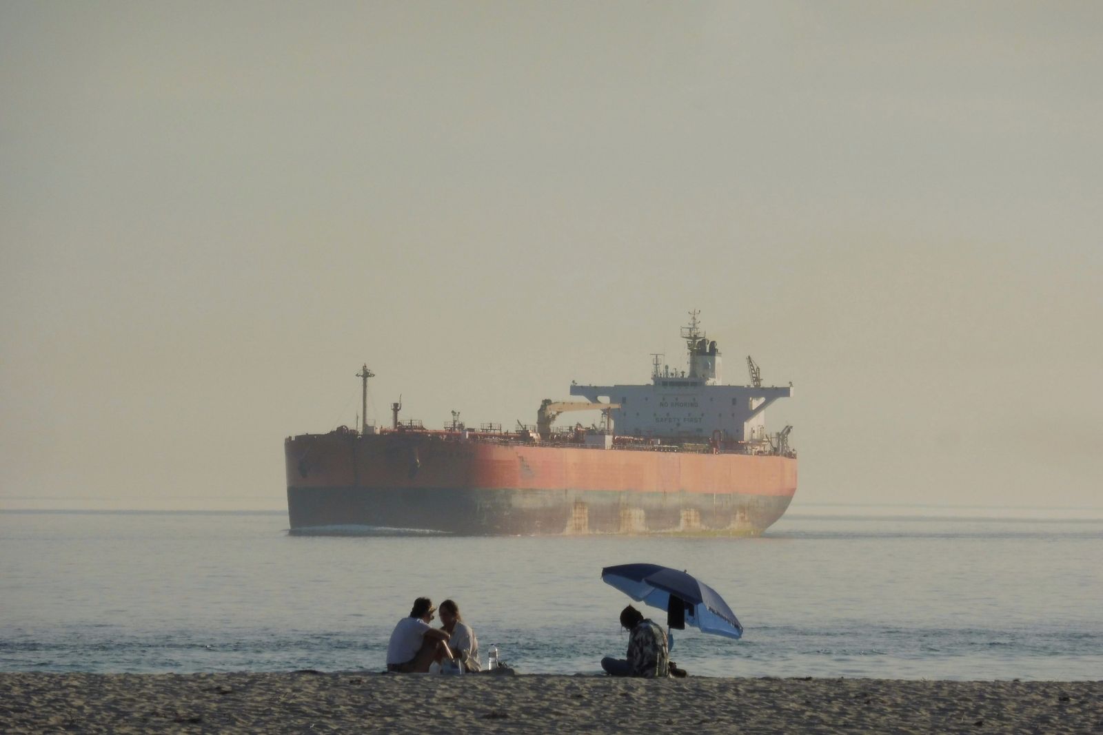 Thick black smoke billows from the hull of a large oil tanker floating in the middle of a calm blue sea.