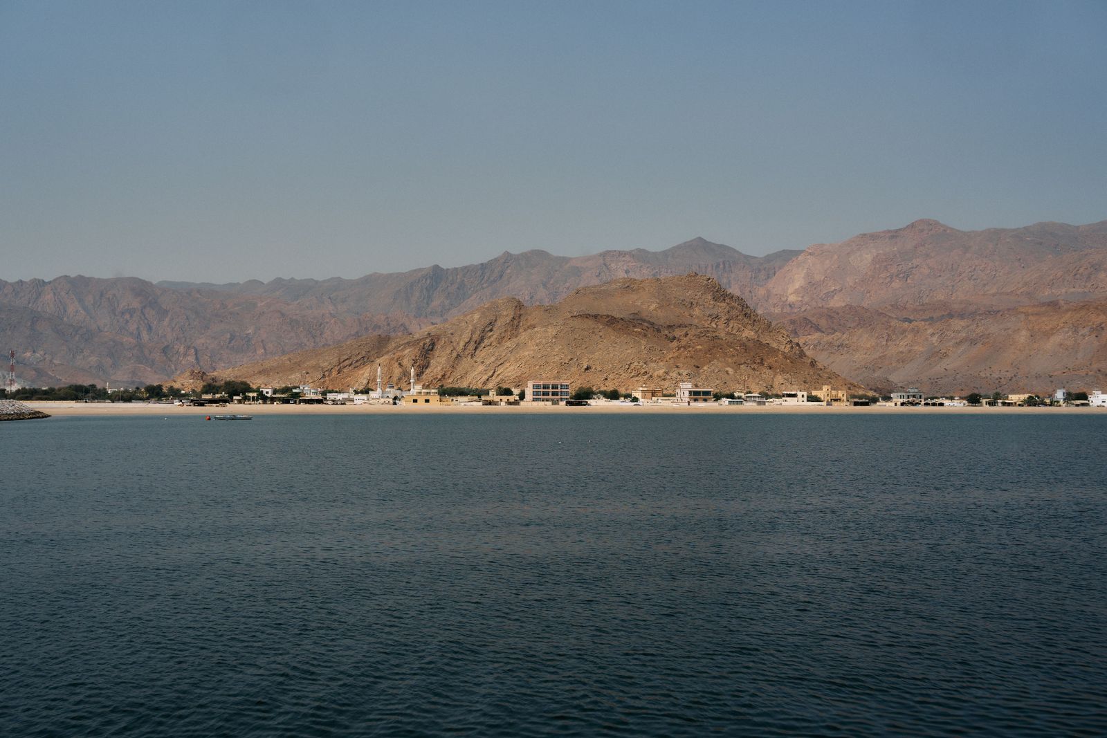 Large container ships navigate the narrow turquoise waters of the Strait of Hormuz between rocky desert coastlines.
