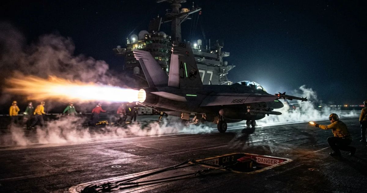 A fighter jet launching from an aircraft carrier at night with a bright afterburner fire.