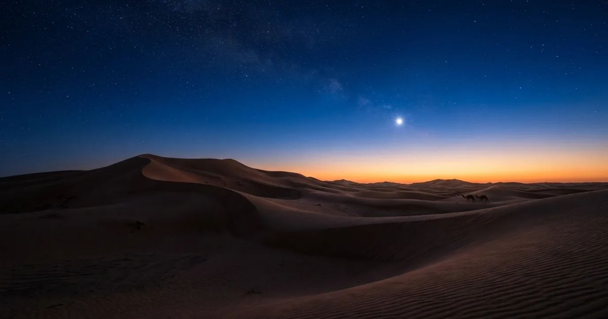 A bright star shines in a dark blue dawn sky above a silhouetted desert landscape with sand dunes.