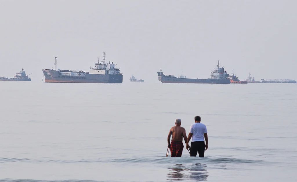 A large oil tanker navigates calm blue waters near a mountainous coastline under a clear, bright sky.